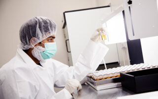 Biologics CMO technician sitting in a lab with a facemask, hair and labcoat visually inspecting vials of drug product