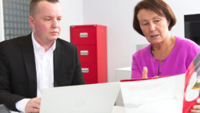 Alison and John sat at a desk with filing cabinets behind them discussing Symbiosis Pharmaceutical Services