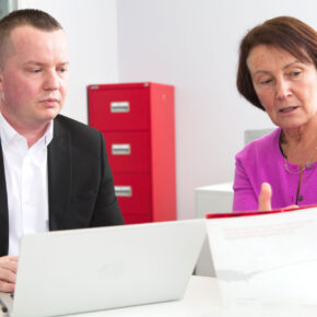 Alison and John sat at a desk with filing cabinets behind them discussing Symbiosis Pharmaceutical Services