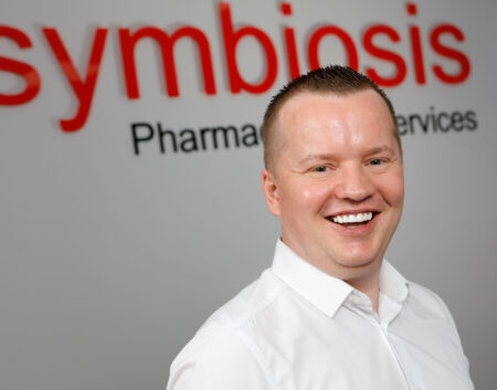 Headshot of John McCormick, member of the Symbiosis leadership team with Symbiosis branded signage behind him