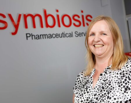 Headshot of Lorraine Den-Kaat with Symbiosis Pharmaceutical Services signage in the background