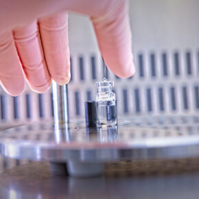 Gloved hand reaching for a drug product vial being filled.