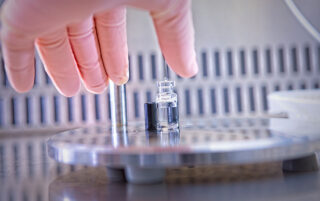 Gloved hand reaching for a drug product vial being filled.