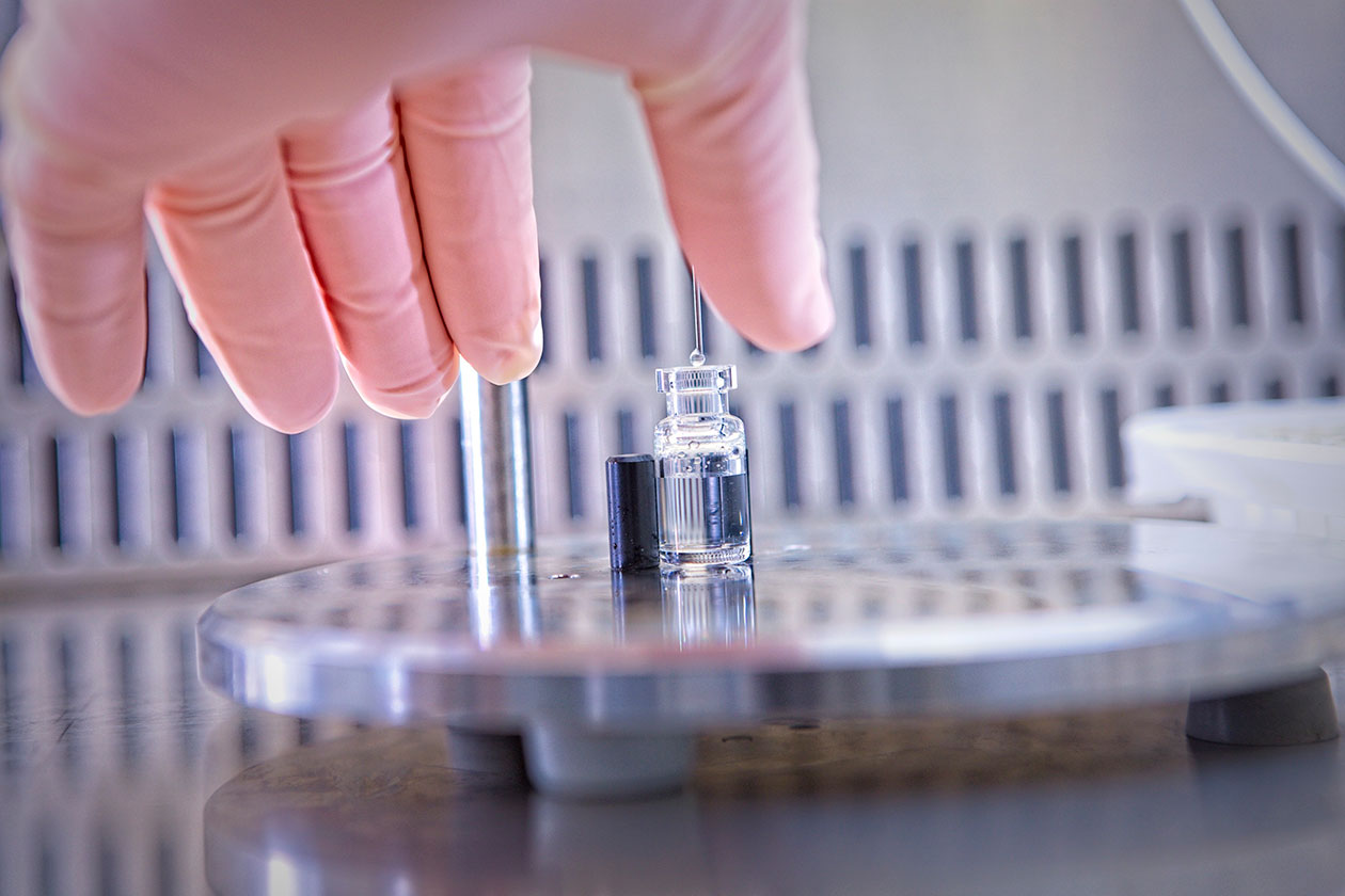 Gloved hand reaching for a drug product vial being filled.